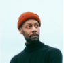 a man in an orange toque and a black turtleneck looks over his shoulder and into the distance