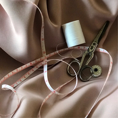 a spool of white thread, a measuring tape and well used metal garment scissors lay atop a blush toned cloth
