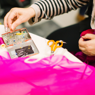 Hands reaching for a ball point pin from a small sewing kit sitting on a white table next to some bright pink tulle. 